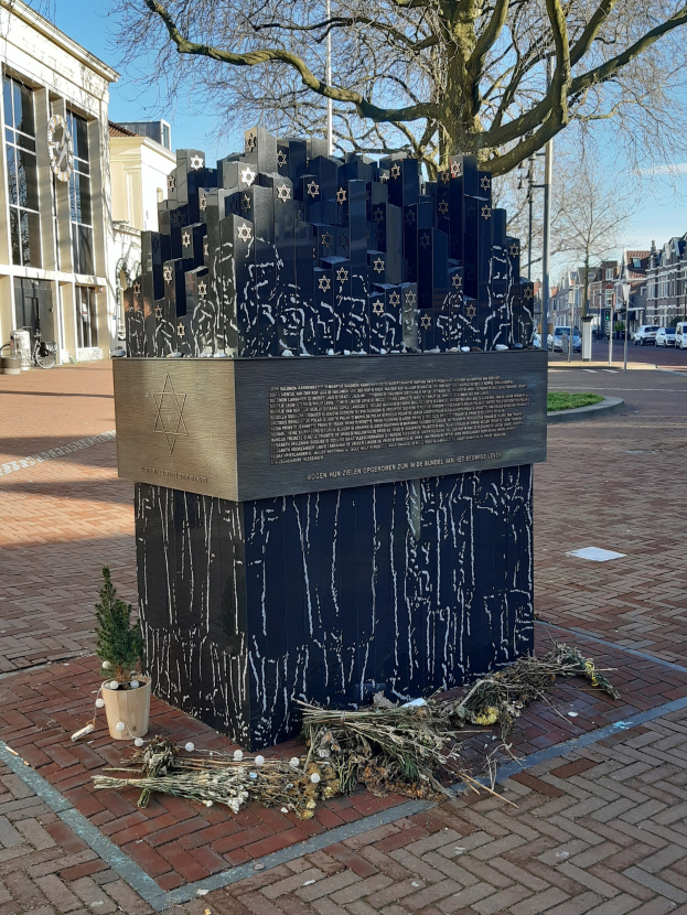 Denkmal in einem Stadtplatz, das Holocaust-Opfern gewidmet ist, mit einem Baum davor, einer Topfpflanze, verstreuten trockenen Blättern und Gebäuden, Fahrzeugen und Bäumen im Hintergrund.