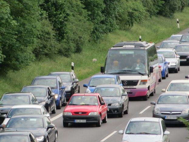 Ein Stau auf einer Autobahn mit vielen Autos und einem Lieferwagen, Menschen in den Fahrzeugen sichtbar, mit Bäumen und Gras im Hintergrund.