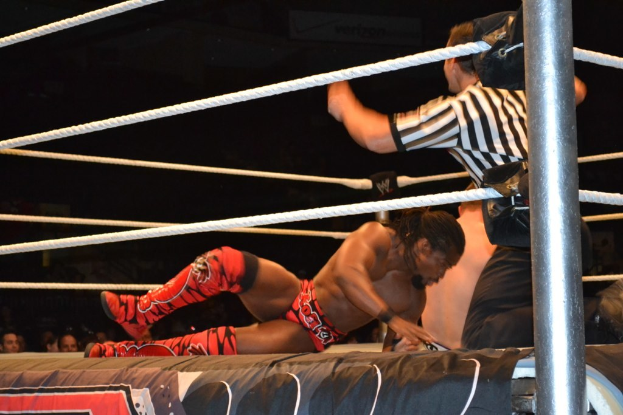 Zwei Männer kämpfen in einem Ring, einer trägt ein rotes Hemd und Schuhe, der andere Shorts, mit einem Schiedsrichter rechts und einer Zuschauermenge im Hintergrund unter einem dunklen Himmel.