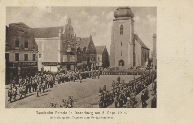 Ein Schwarz-Weiß-Foto eines Umzugs in Insterburg am 5. September 1914 mit vielen Menschen und Gebäuden im Hintergrund und einem Text am unteren Bildrand.