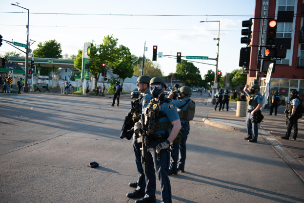Eine Gruppe von Polizisten in Helmen und Uniformen steht auf einer Straße und hält Waffen, mit Fahrzeugen, Verkehrszeichen, Bäumen, Gebäuden und einem bewölkten Himmel im Hintergrund.