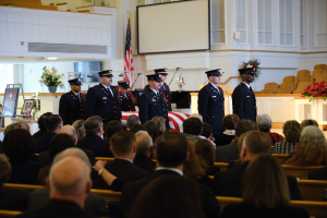 Eine Gruppe uniformierter Personen mit Mützen steht vor einem sitzenden Publikum bei einer Trauerfeier, mit Blumensträußen, einer Fahne, einem Bildschirm und einer Wand im Hintergrund.
