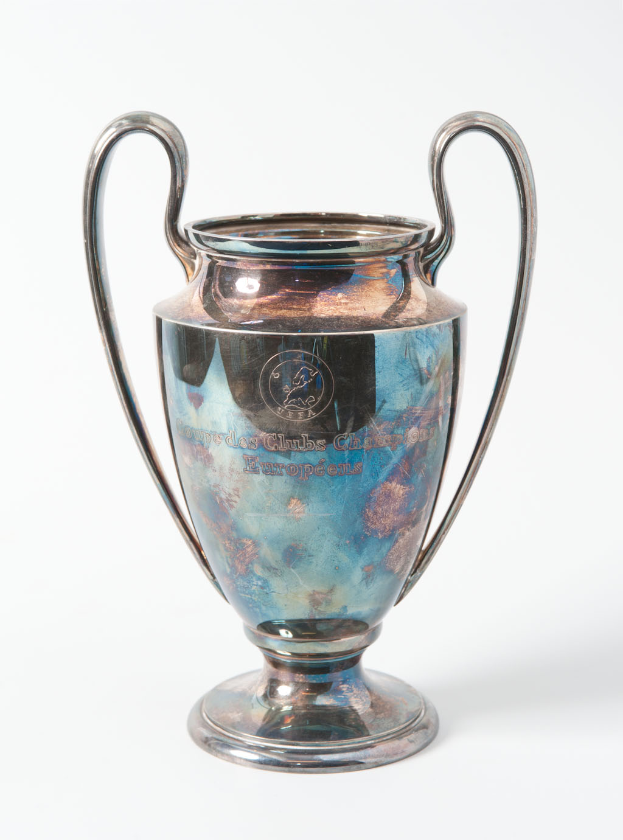 Der UEFA Champions League Pokal, ein stählerner Becher mit zwei Griffen und einem blauen und silbernen Farbschema, mit der Inschrift "UEFA Champions League", vor einem weißen Hintergrund.