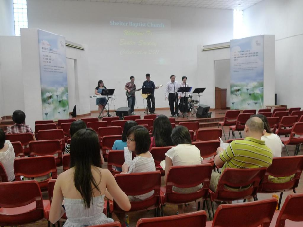 Eine Auditoriumsbühne mit zwei Gitarristen in der Mitte und anderen, die singen und Instrumente spielen, in den Ecken, mit Bannern neben der Bühne und einem Publikum davor, das Papiere und Bücher hält.