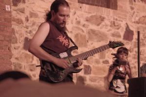 Ein bärtiger Mann spielt Gitarre auf der Bühne, steht und zupft die Saiten, während eine Frau mit Mikrofon rechts von ihm steht, mit einer Menge von Menschen im Vordergrund und einer Wand mit einem Fenster im Hintergrund.