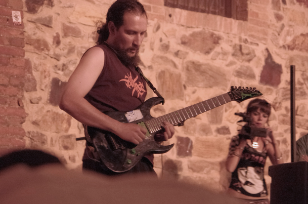 Ein bärtiger Mann spielt Gitarre auf der Bühne, steht und zupft die Saiten, während eine Frau mit Mikrofon rechts von ihm steht, mit einer Menge von Menschen im Vordergrund und einer Wand mit einem Fenster im Hintergrund.