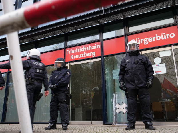 Polizeibeamte in Helmen und Handschuhen vor einem Glaswandgebäude mit angeklebten Papieren, einem Pfahl links und Schildern mit der Aufschrift "Friedrichshain Kreuzberg" im Hintergrund.