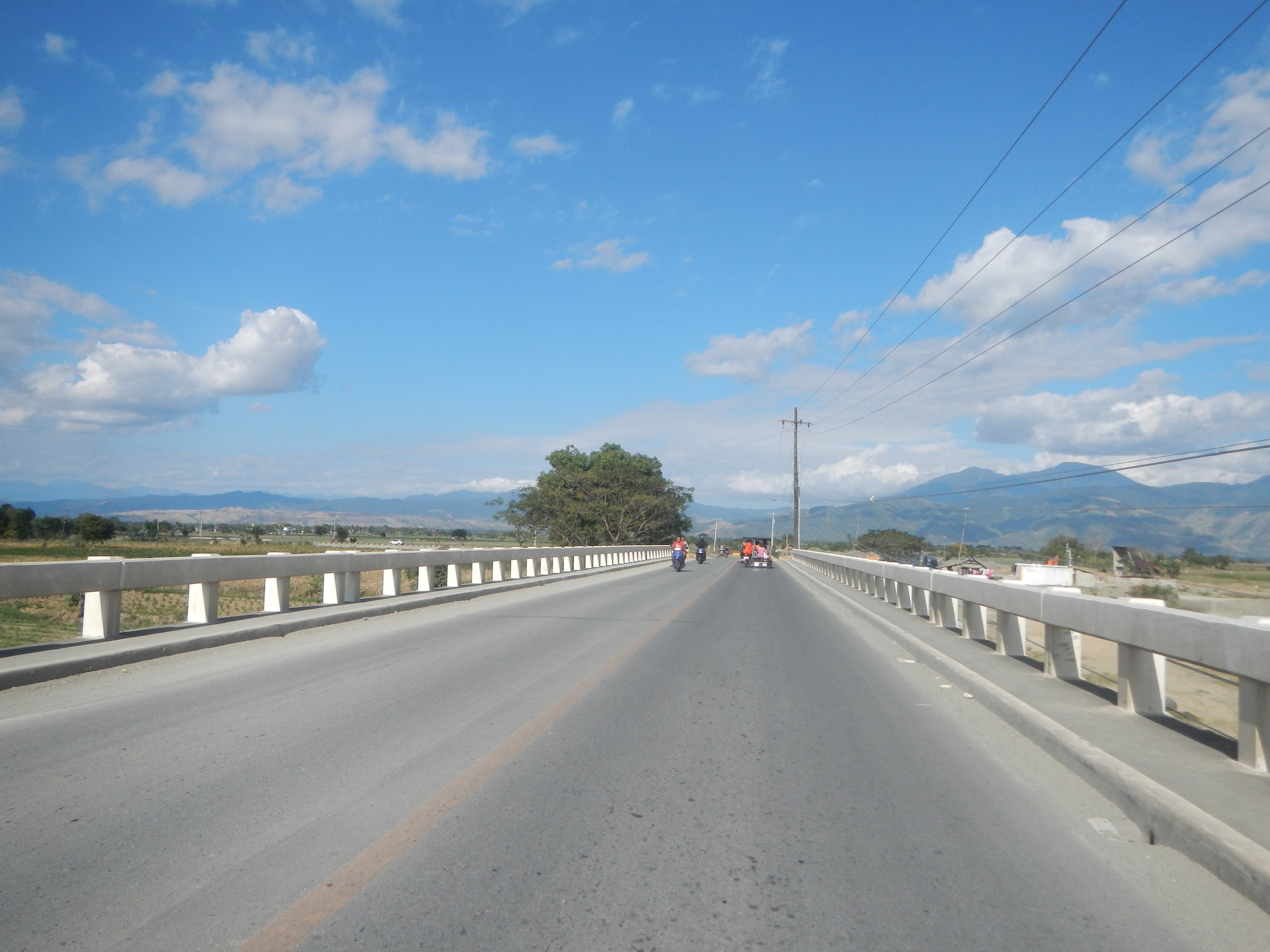 Autobahn mit Motorradfahrern, Geländern, Strommasten, Bäumen, Hügeln und einem bewölkten Himmel im Hintergrund.