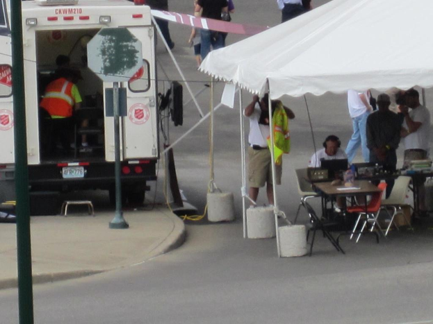 Ein Rettungswagen steht links von der Straße, während rechts ein Zelt viele Menschen schützt, mit einem Tisch und einem Stuhl in der Nähe.