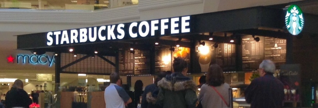 Menschen vor einem Coffeeshop mit einem Namensschild, Logo, Menü und Lampen, mit einem weiteren Geschäft daneben und einem Gang darüber.