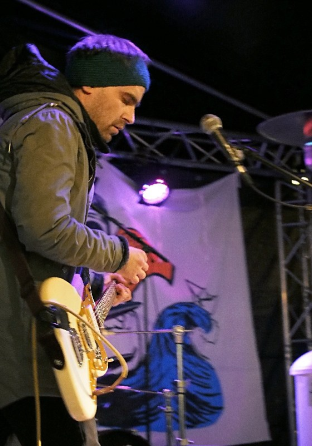 Ein Mann in einer Jacke und einer Mütze spielt eine E-Gitarre auf der Bühne, mit einem Mikrofon davor, einem Schlagzeug rechts von ihm, Scheinwerfern und einem Banner im Hintergrund.
