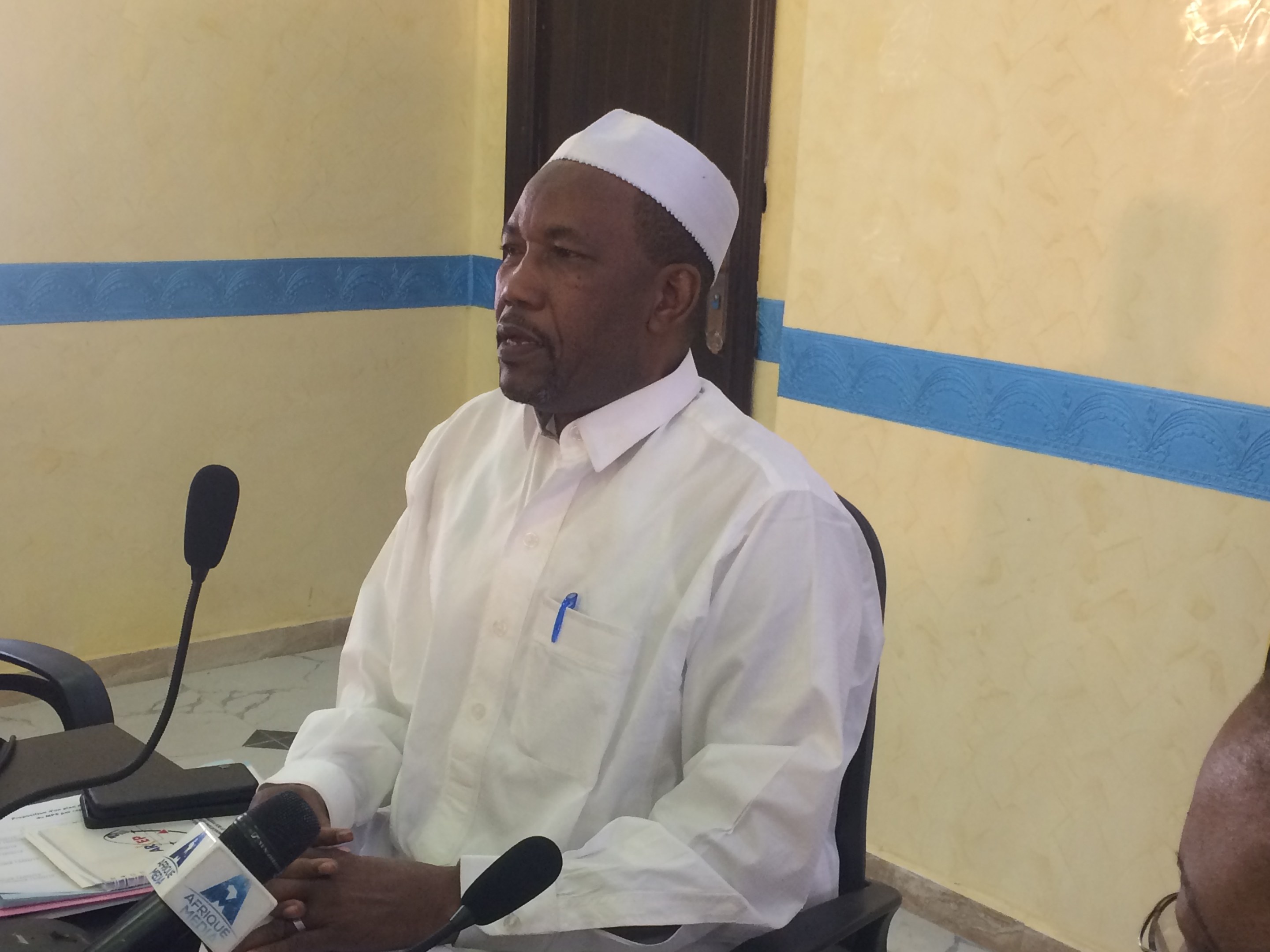 Ein Mann in weißem Anzug und Hut sitzt an einem Tisch mit Mikrofonen und spricht, während Papiere und Gegenstände auf dem Tisch zu sehen sind; rechts von ihm sitzt eine Person mit Brille, im Hintergrund eine Tür und eine Wand.