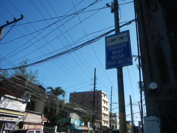 Stadtstraße mit fahrenden Autos, Strommäste mit Drähten, Gebäude, Bäume und ein Schild an einem Pfahl mit der Aufschrift "Fahrer müssen Sitzgürtel tragen."