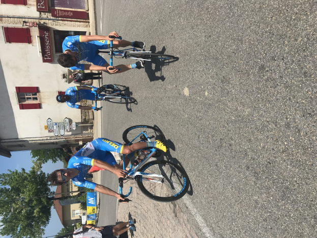 Eine Gruppe von Radfahrern fährt eine Straße entlang an einem Gebäude mit Fenstern und Schildern mit Text vorbei, mit Bäumen und einem blauen Himmel im Hintergrund und Zuschauern auf der Straße darunter.