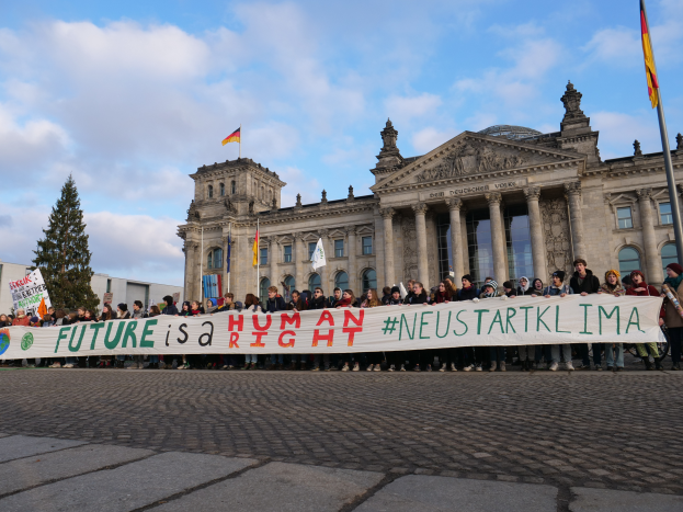 Eine Gruppe von Menschen steht vor dem Reichstaggebäude in Berlin und hält eine Fahne mit der Aufschrift "Zukunft ist ein Menschenrecht", während das Gebäude mit Säulen, Fenstern und Bögen verziert ist und von Bäumen und Fahnen umgeben ist, unter einem bewölkten Himmel.