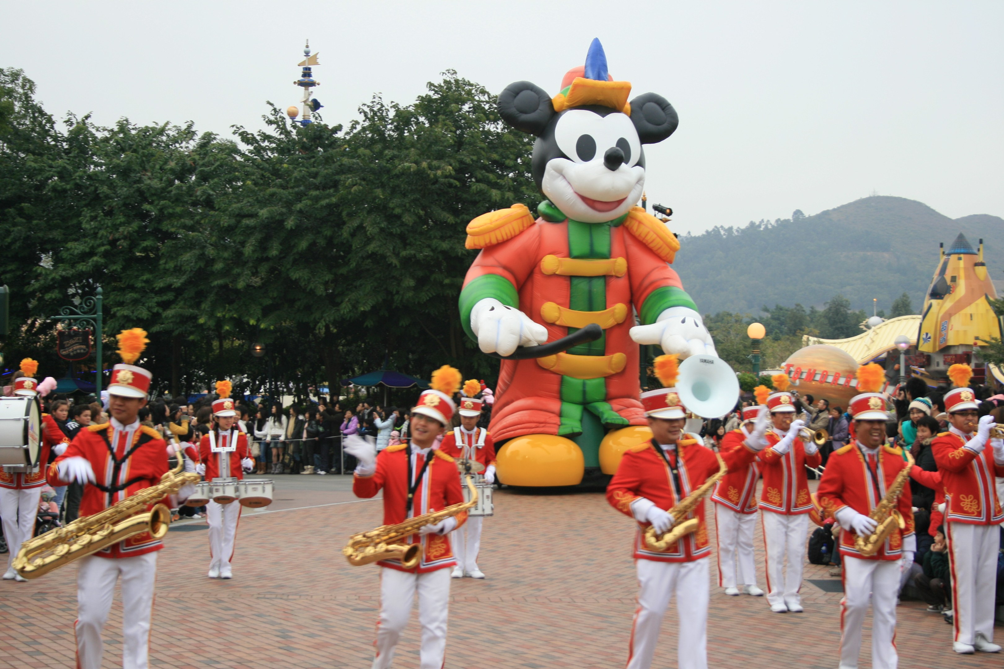 Eine Gruppe von Menschen in Kostümen spielt Musikinstrumente neben einem Mickey-Maus-Floß bei einem Umzug, mit Bäumen, Gebäuden und einem klaren Himmel im Hintergrund; ein Schild mit Text ist links sichtbar.