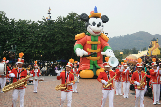 Eine Gruppe von Menschen in Kostümen spielt Musikinstrumente neben einem Mickey-Maus-Floß bei einem Umzug, mit Bäumen, Gebäuden und einem klaren Himmel im Hintergrund; ein Schild mit Text ist links sichtbar.