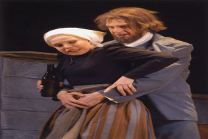 Ein Mann im Anzug mit ernstem Gesichtsausdruck und eine Frau mit einer Flasche stehen auf einer Bühne mit Text unten und einer Wand im Hintergrund.