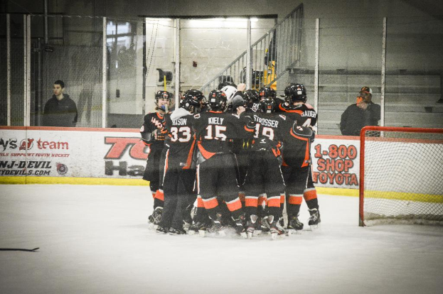 Eine Gruppe von Menschen in Schlittschuhen, Handschuhen und Helmen steht auf einem Eisfläche mit Absperrungen und Glastüren dahinter, einem Hockey-Tor mit Netz auf der rechten Seite und einem Hockey-Schläger auf der linken Seite.