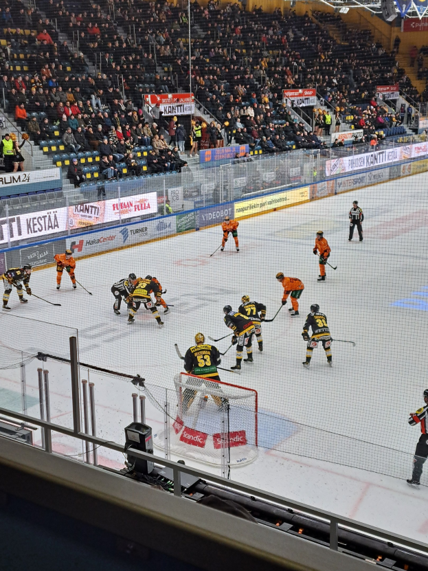 Hockeyspieler bei einem Spiel auf einer Indoor-Eisfläche mit Netz und Umzäunung, Zuschauern im Hintergrund und beleuchteter Stadionatmosphäre.