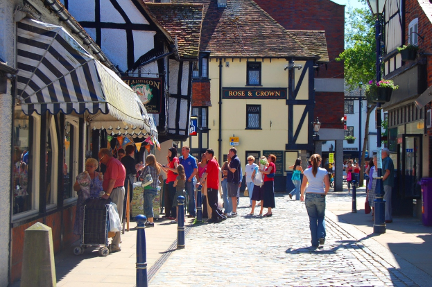 Eine Straße mit Pfählen und Gebäuden auf beiden Seiten, Menschen auf der linken Seite mit einem Handwagen, ein Mann und eine Frau auf der rechten Seite in der Nähe eines violetten Müllcontainers, mit Gebäuden, Bäumen und Blumentöpfen im Hintergrund.