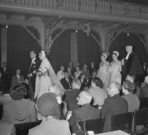 Schwarz-weiß-Foto einer Hochzeitszeremonie mit Gästen auf Stühlen und Stehenden in einem großen Raum, Tische mit Tassen, Sträußen, Vorhängen und Deckenleuchten im Hintergrund.