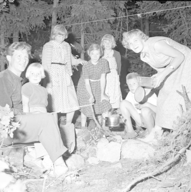 Ein Schwarz-Weiß-Foto einer Familie, die sich um ein Lagerfeuer im Wald versammelt hat, mit einigen sitzend und einigen stehend, einer Person, die ein Objekt hält, umgeben von Bäumen, einem Draht und einem Boden, der mit Steinen und Pflanzen bedeckt ist.
