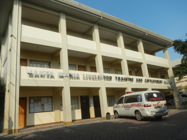 Ein zweistöckiges Gebäude namens Santa Maria Lebensmittel-Trainings- und Beschäftigungszentrum mit Fenstern, Türen und Beschriftung, umgeben von Bäumen und Fahrzeugen auf einer Straße unter einem klaren blauen Himmel.