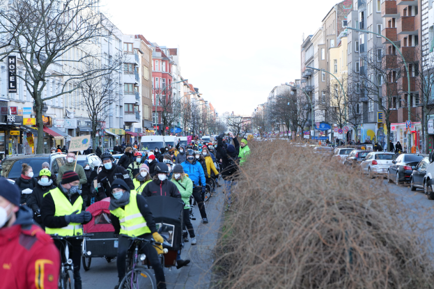 Eine große Gruppe von Menschen mit Masken und Sicherheitswesten fährt auf Fahrrädern eine straße mit Bäumen, Gebäuden, Laternen und Texttafeln entlang, während Fahrzeuge die Straße teilen und trockenes Gras die rechte Seite unter einem klaren blauen Himmel einnimmt.
