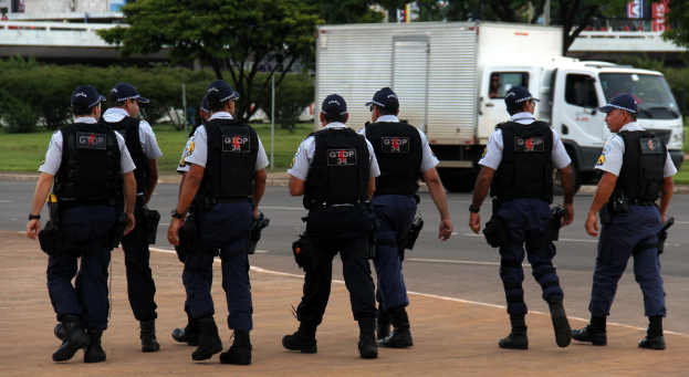 Eine Gruppe von Beamten in Uniform, einschließlich Mützen, Hemden, Schutzwesten, Uhren, Hosen und Schuhen, die auf einer Straße mit einem rechts geparkten Lkw in der Nähe eines Zauns, Gebäuden, Bäumen, Pflanzen, Polen und Bannern im Hintergrund und einer Brücke im oberen rechten Eck gehen.