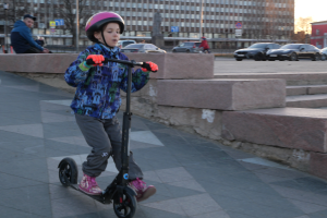 Ein junger Junge fährt mit einem Roller auf einem Gehweg, trägt einen Helm und Handschuhe, mit Stufen, Fahrzeugen, Menschen, Bäumen, Pfosten, Brettern, Gebäuden und einem klaren blauen Himmel im Hintergrund.