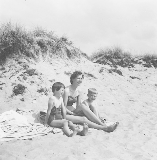 Schwarz-weiß-Foto einer Frau in einem Bikini und zwei Kindern in Badebekleidung, die auf einer Decke am Strand sitzen, mit Sand, Gras und Himmel im Hintergrund.