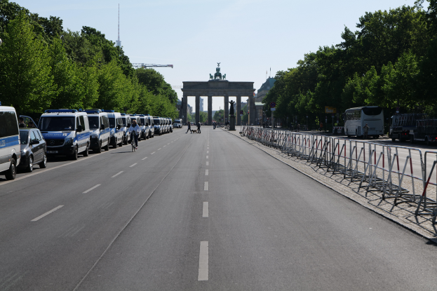 Eine Reihe von Polizeifahrzeugen auf einer Straße vor dem Brandenburger Tor in Berlin, Deutschland, mit Menschen auf Fahrrädern und Stehenden, Barrieren, Bäumen und einem Bogen mit Statuen im Hintergrund.