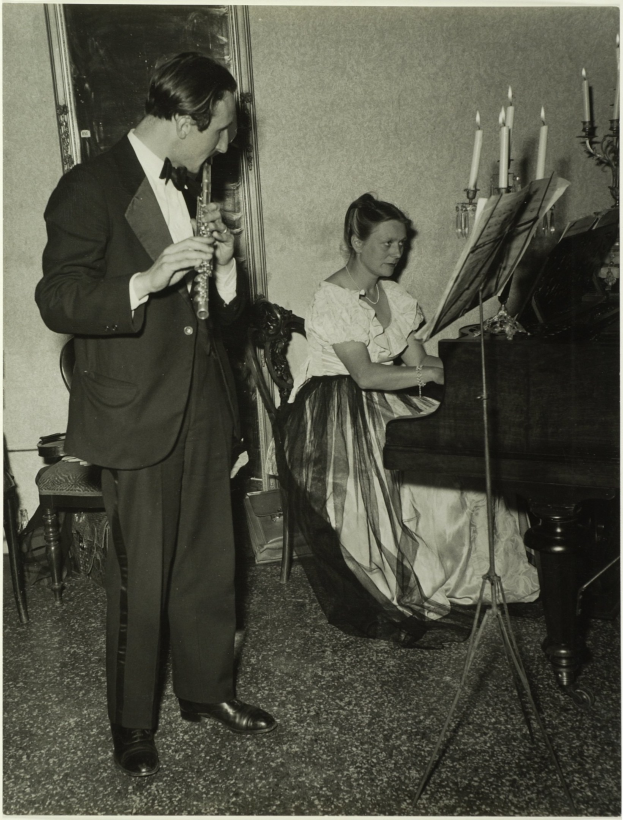 Ein Mann im Smoking spielt Klarinette, während er neben einer Frau steht, die auf einem Piano sitzt und ein Buch auf dem Notenständer hat, mit einem gerahmten Bild an der Wand dahinter.