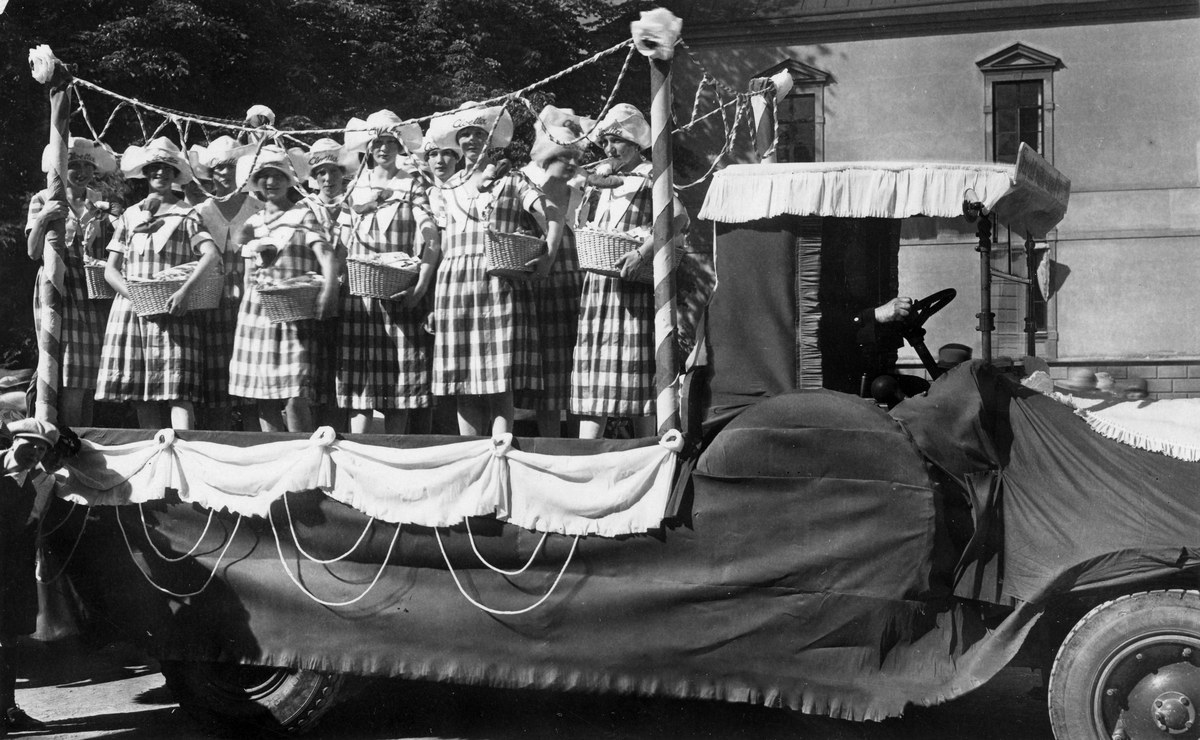 Schwarzes und weißes Foto einer Gruppe von Frauen, die auf einem Lastwagen stehen und Körbe halten, mit einem Gebäude und Bäumen im Hintergrund.