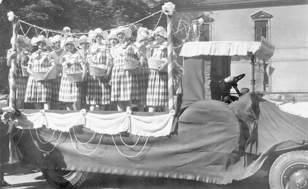Schwarzes und weißes Foto einer Gruppe von Frauen, die auf einem Lastwagen stehen und Körbe halten, mit einem Gebäude und Bäumen im Hintergrund.