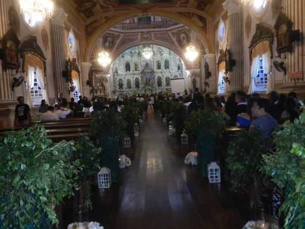 Eine Hochzeitszeremonie in einer Kirche mit Gästen auf Bänken und im Gang, dekoriert mit Zimmerpflanzen, Skulpturen an den Wänden, Vorhängen an den Fenstern und hängenden Kronleuchtern, mit einem Bildschirm im Hintergrund.