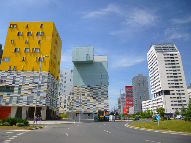Eine Stadtstraße mit hohen Gebäuden im Hintergrund, Straßenlaternen, Autos, Informationsschildern, Büschen, Bäumen und einem bewölkten Himmel, mit den einzigartig gestalteten Gebäuden der Universität Rotterdam.