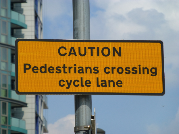 Gelbes Warnschild an einem Pfahl vor einem Gebäude mit einem klaren blauen Himmel im Hintergrund, auf dem steht "Fußgänger überqueren Fahrradspur".
