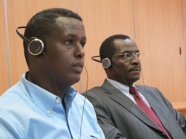 Zwei Männer in einem Gerichtssaal mit Kopfhörern, einer in einem blauen Hemd links und einer mit Brille in der Mitte, vor einer hölzernen Wand sitzend.