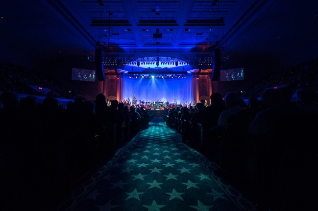 Ein großer Zuschauerraum mit Sitzplätzen, eine Bühne mit Musikern und Deckenbeleuchtung und Bildschirme, was auf ein Konzerthaus während der Nationalkonvention 2019 hinweist.