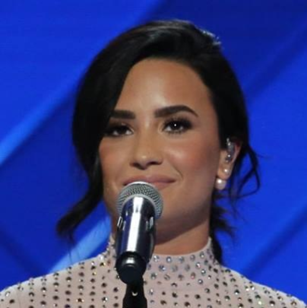 Demi Lovato steht selbstbewusst bei den Grammys mit einem eleganten Bob und einem schwarzen Kleid vor einem Mikrofon auf einem blauen Hintergrund.