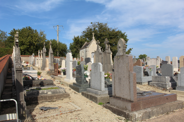 Ein Friedhof mit zahlreichen Grabsteinen und Blumen im Vordergrund, ein Gebäude im Hintergrund, Bäume, ein Strommast mit Kabeln und ein bewölkter Himmel.
