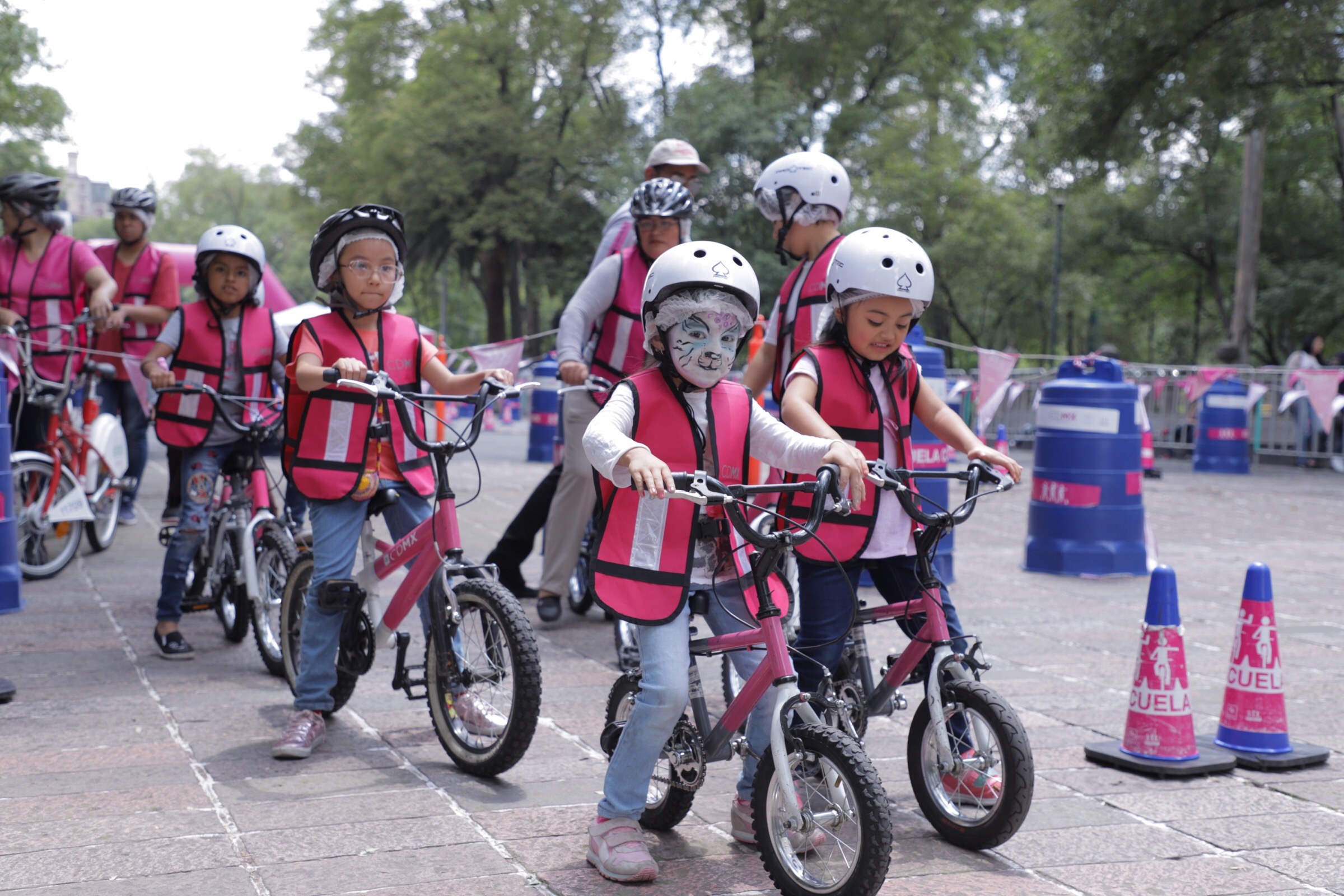 Kinder mit Helmen fahren Fahrräder auf einer Straße mit Verkehrsmarkierungen, einige mit Gesichtsbemalung, vor einem Hintergrund aus Bäumen und einem klaren Himmel.
