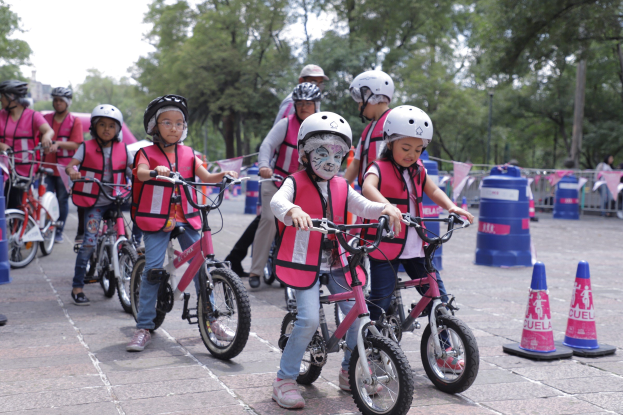 Kinder mit Helmen fahren Fahrräder auf einer Straße mit Verkehrsmarkierungen, einige mit Gesichtsbemalung, vor einem Hintergrund aus Bäumen und einem klaren Himmel.