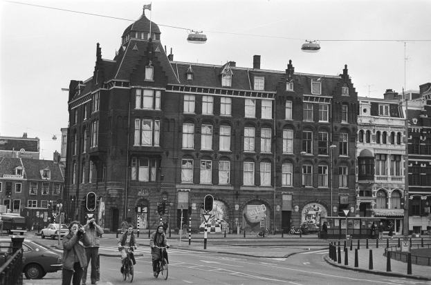 Ein Schwarz-Wei├č-Foto von Menschen, die auf der Stra├če Fahrr├Ąder fahren, umgeben von Geb├Ąuden, Stra├čenlaternen, Verkehrspf├Ąlen, Verkehrsampeln, Kraftfahrzeugen, Passanten, Absperrpfeilern, Stromkabeln und Himmel.
