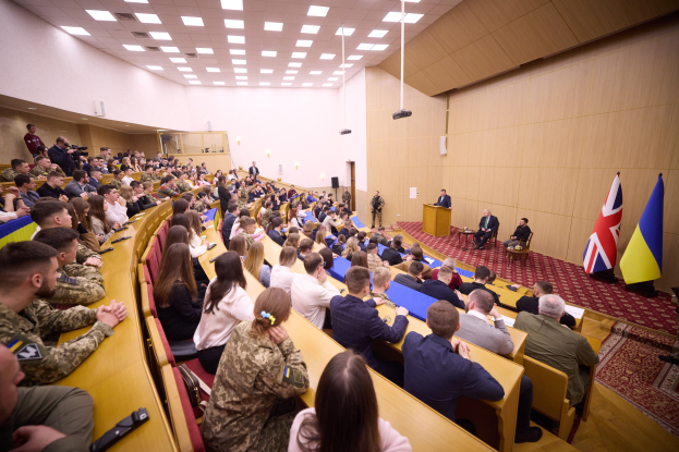 Eine große Gruppe von Menschen an Tischen in einem Raum mit Flaggen auf der rechten Seite, Papiere und Gegenstände auf den Tischen, einigestehend in der Nähe eines Podiums mit einem Mikrofon und Deckenleuchten und ein Projektor, der eine militärische Schulungseinrichtung andeutet.