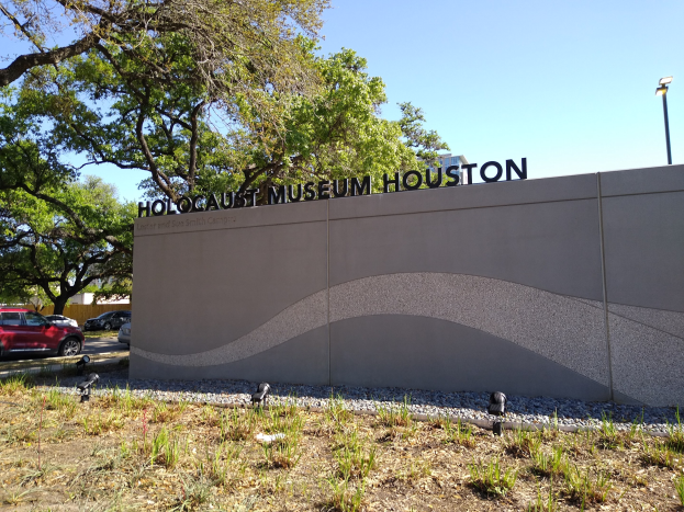 Außenansicht des Houston Holocaust Museums mit einer Gedenkwand, Steinen, Grünfläche, Bäumen, vorbeifahrenden Fahrzeugen, einem Straßenpfahl und einem bewölkten Himmel.