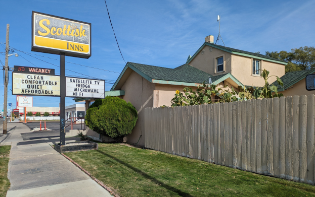 Außenansicht eines schottischen Motels in San Francisco, Kalifornien, das ein Haus mit Dach und Fenstern, einen Holzzaun, Pflanzen, Gras, Bäume, Schilder, Strommasten mit Drähten, eine Straßenlaterne, Fahrzeuge auf der Straße und einen bewölkten Himmel zeigt.