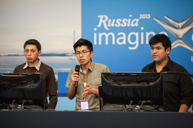 Drei Jungen stehen neben einem Schreibtisch mit zwei Computern, einer spricht in ein Mikrofon, trägt ID-Karten mit roten Markierungen, vor einer Wand mit einem Werbeplakat, auf dem "Russia Imagine 2013" steht.